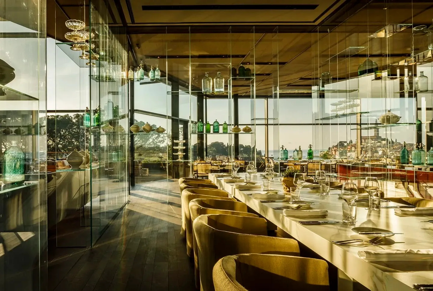 Long communal chef's table in a glass-walled restaurant overlooking Rovinj at golden hour